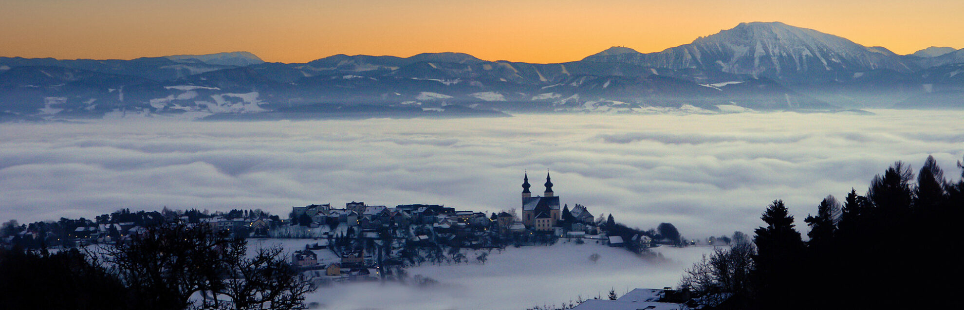 Winterliche Landschaft in Österreich mit verschneitem Alpenpanorama und malerischem Bergdorf im Nebelmeer – ein Highlight für Busreisen in die Alpen.