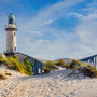 Leuchtturm hinter Sanddüne und modernem Gebäude bei sonnigem Wetter.