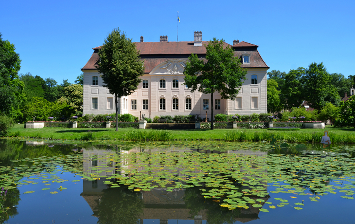 Herrenhaus mit Spiegelung in einem Teich voller Seerosen, sonniges Wetter.