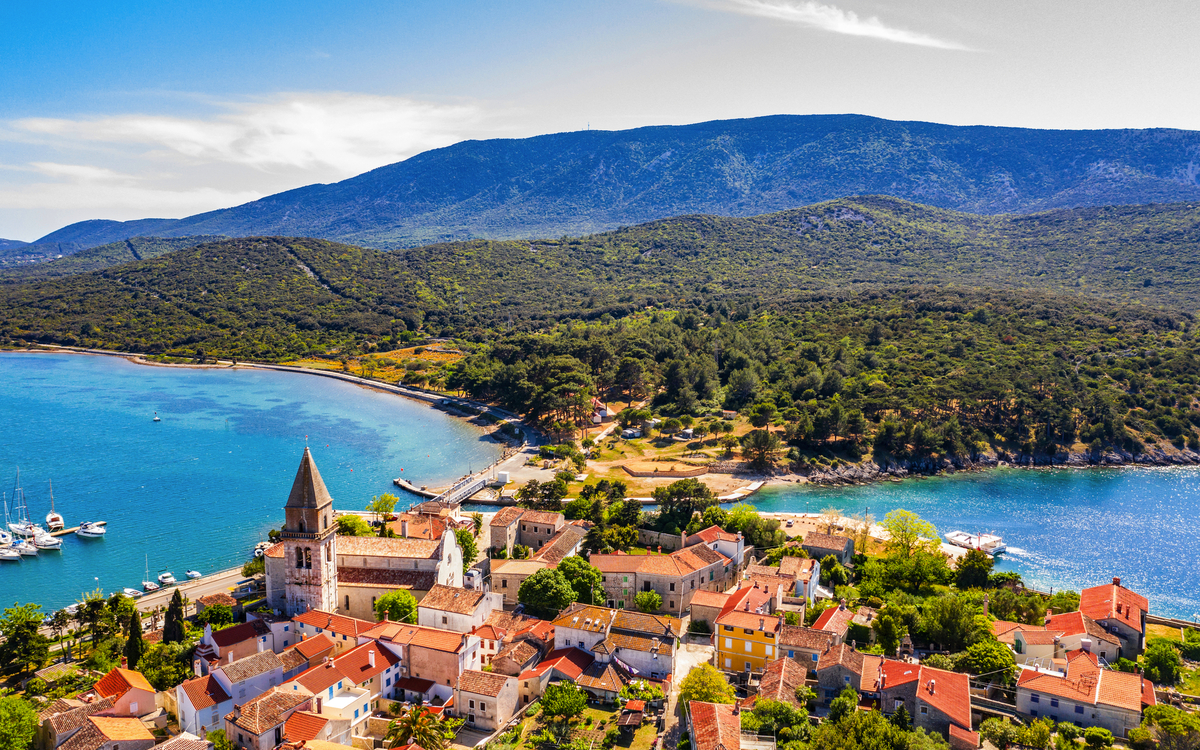 Luftaufnahme einer Küstenstadt mit Kirche, Meer und Bergen im Hintergrund.