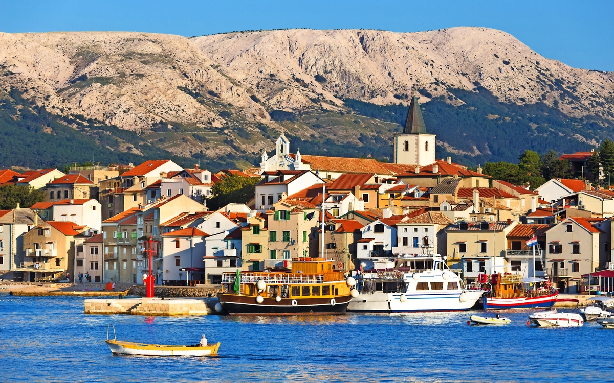 Küstenort mit malerischen Gebäuden, Kirche und Booten im Hafen, im Hintergrund bergige Landschaft.