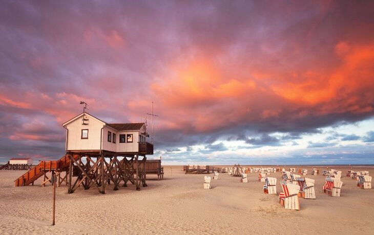 Haus auf Stelzen am Strand bei Sonnenuntergang mit Wolken und Strandkörben.