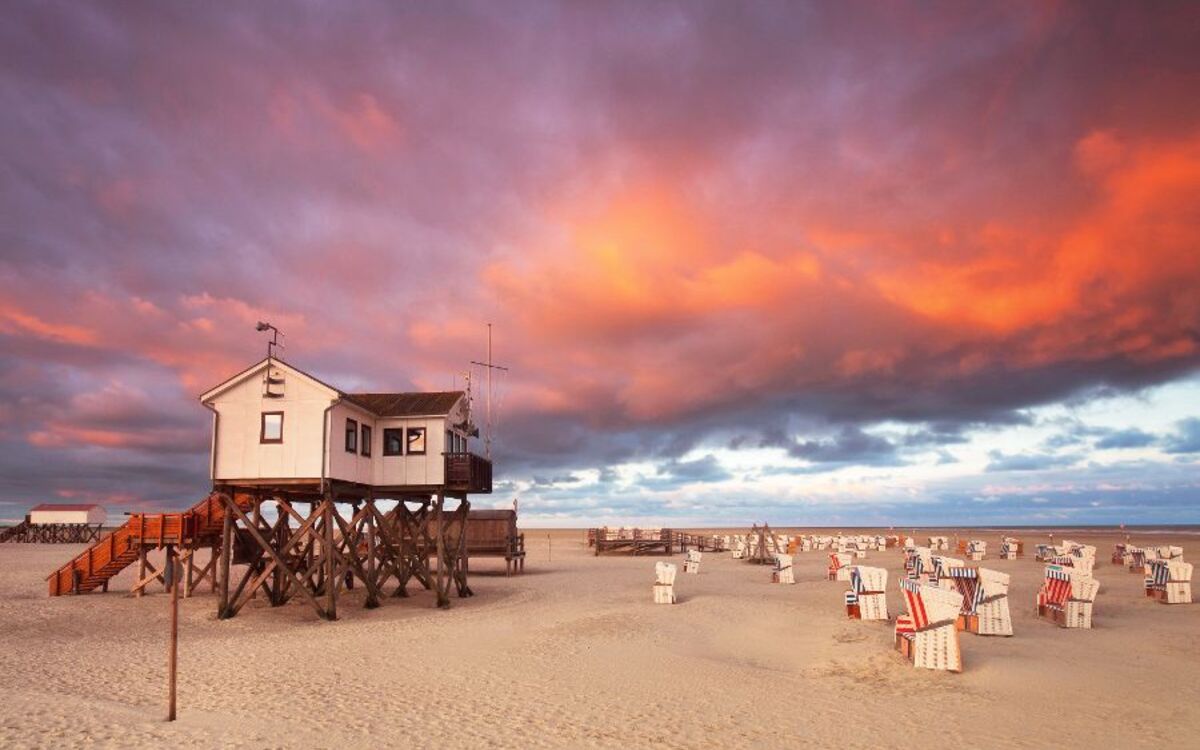 Haus auf Stelzen am Strand bei Sonnenuntergang mit Wolken und Strandkörben.