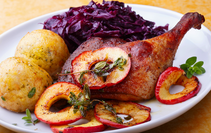 Gebackene Entenkeule mit Rotkohl, Klößen und Apfelscheiben auf einem weißen Teller.