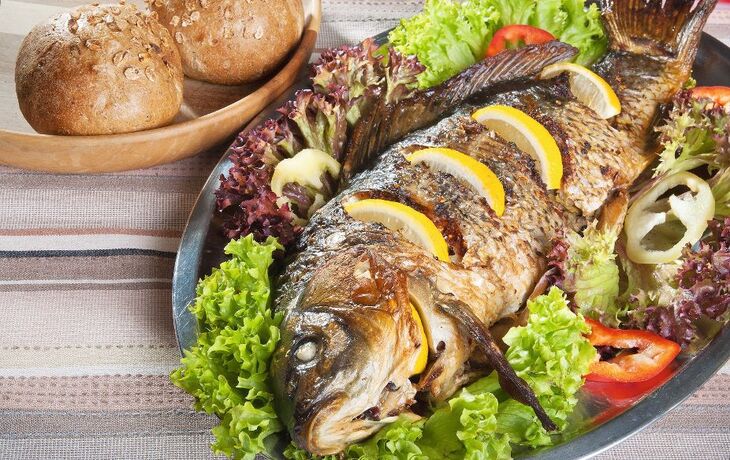 Gegrillter Karpfen auf Salatbett mit Zitronenscheiben und Brötchen im Hintergrund.