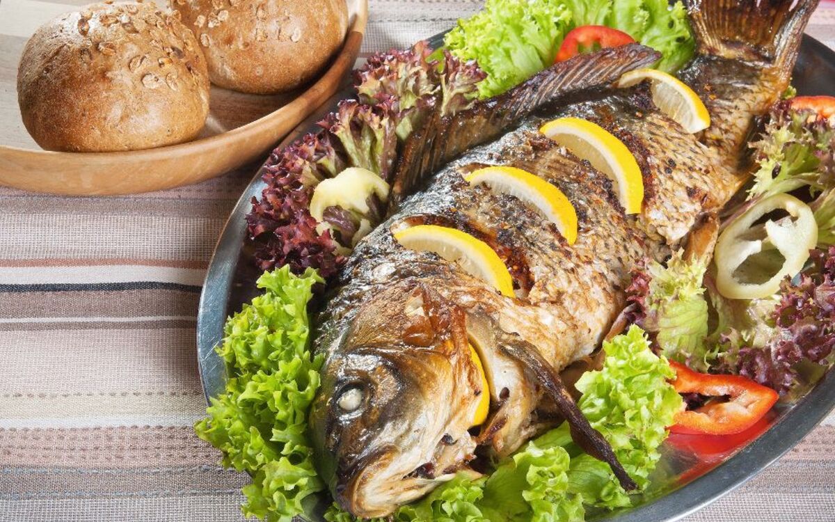 Gegrillter Karpfen auf Salatbett mit Zitronenscheiben und Brötchen im Hintergrund.