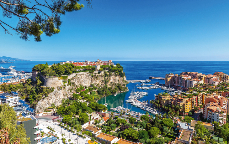 Blick auf eine Küstenstadt mit einer felsigen Halbinsel, auf der sich eine Festung befindet. Im Vordergrund sind Grünflächen und Gebäude sichtbar, dahinter ein Yachthafen mit zahlreichen Booten. Im Hintergrund erstreckt sich das blaue Meer unter einem klaren Himmel.