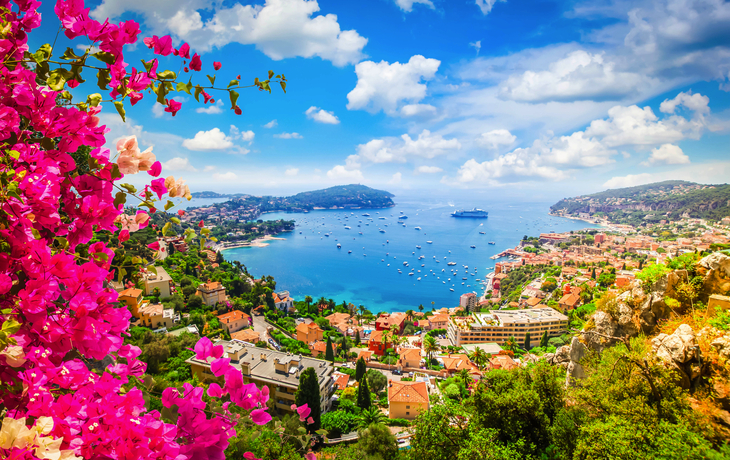 Blick auf die Mittelmeerküste von Nizza an der Côte d'Azur, mit blauem Wasser und der Promenade im Vordergrund, umgeben von üppiger Vegetation und einer lebhaften Stadtansicht.