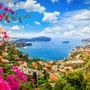 Blick auf die Mittelmeerküste von Nizza an der Côte d'Azur, mit blauem Wasser und der Promenade im Vordergrund, umgeben von üppiger Vegetation und einer lebhaften Stadtansicht.