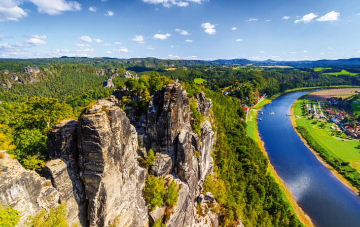 Felsformationen in grünem Tal mit Fluss und blauem Himmel. Ansicht auf Rathen von der Bateibrücke in der Sächsischen Schweiz, Deutschland
