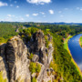 Felsformationen in grünem Tal mit Fluss und blauem Himmel. Ansicht auf Rathen von der Bateibrücke in der Sächsischen Schweiz, Deutschland