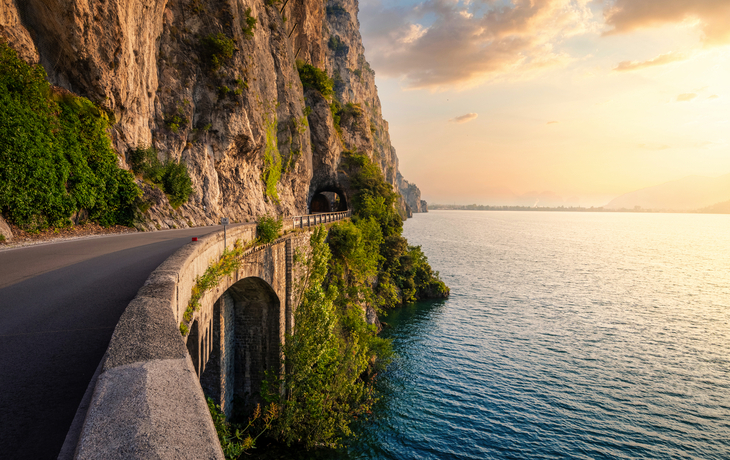 Küstenstraße entlang felsiger Klippe und See im Sonnenuntergang.