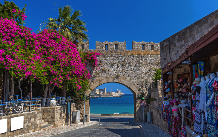Stadttor mit Bougainvillea, Meerblick und Souvenirladen im Vordergrund.
