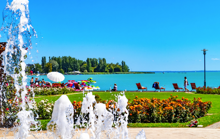Brunnen vor Strandpromenade mit Spaziergängern und Liegestühlen am Wasser.