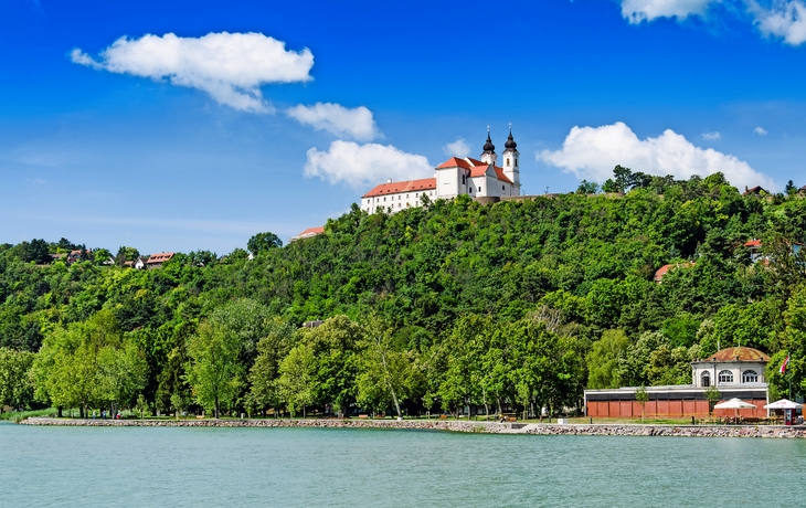 Kloster auf einem Hügel mit Bäumen und blauem Himmel im Hintergrund.