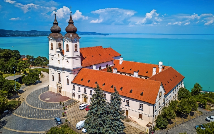 Luftaufnahme einer Kirche mit zwei Türmen am Ufer eines blauen Sees.