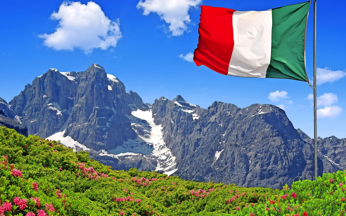 Italienische Flagge weht vor Bergkulisse mit Blumenwiese.