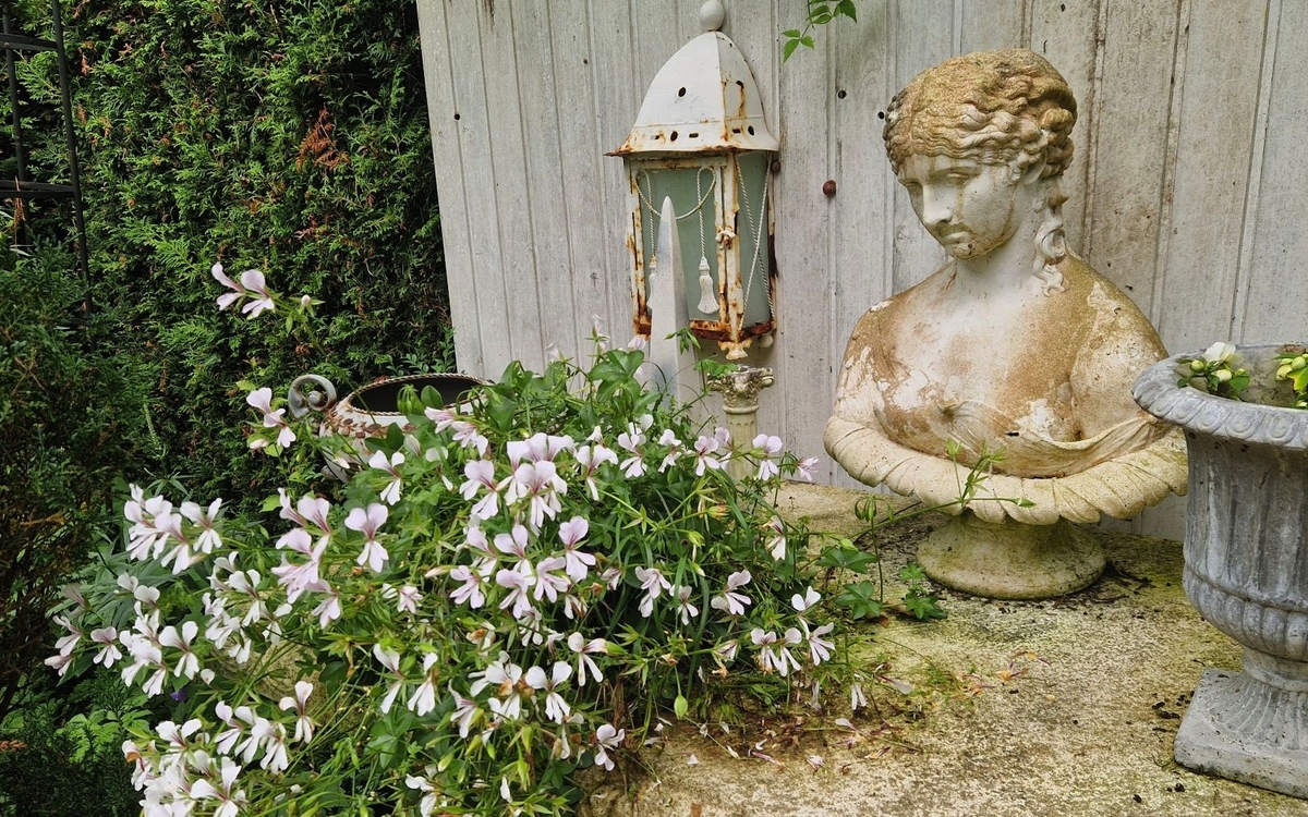 Büste und Laterne im Garten neben blühenden Pflanzen und einem Blumentopf.