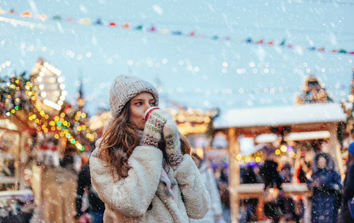 Frau mit Mütze und Handschuhen trinkt auf einem Weihnachtsmarkt im Schnee.