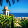 Glockenturm vor der Kulisse von Sanremo an der ligurischen Küste mit Blick auf das Meer und Palmen.
