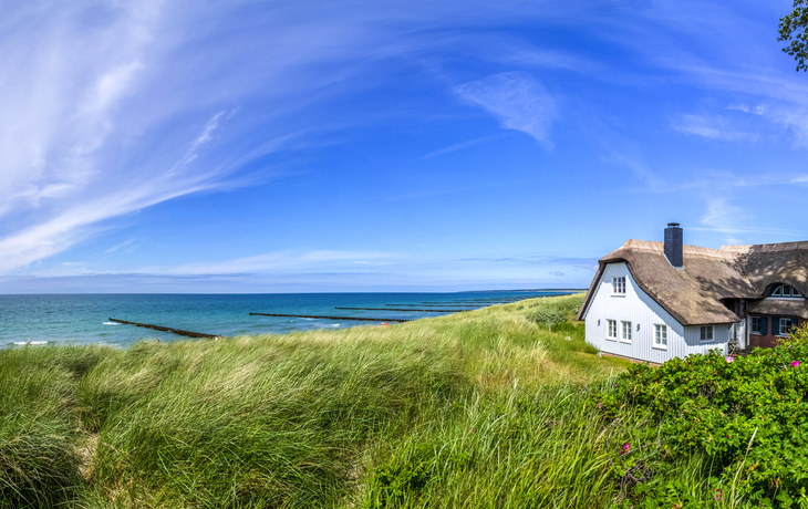 Reetdachhaus am Strand mit blauem Himmel und Dünenvegetation.