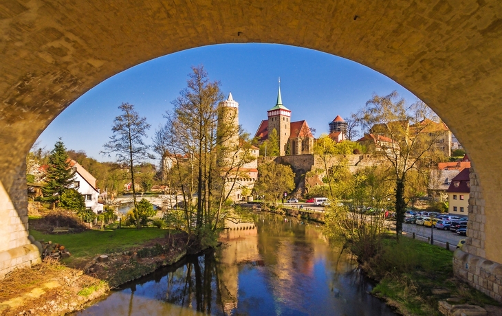 Blick von unter Brücke auf Fluss und Stadt im Sommer mit Türmen und Bäumen.