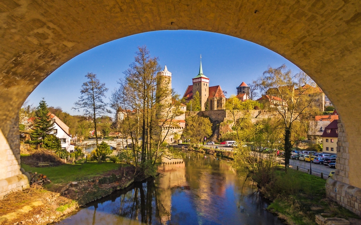 Blick von unter Brücke auf Fluss und Stadt im Sommer mit Türmen und Bäumen.