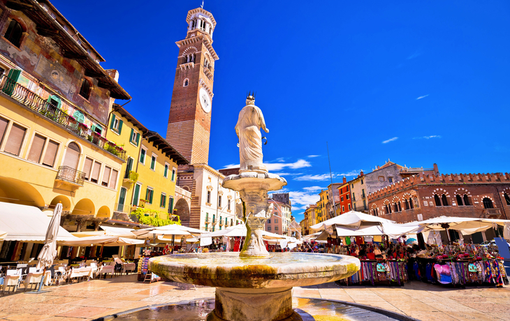 Marktplatz mit Brunnen und Uhrturm an einem sonnigen Tag in einer europäischen Stadt.
