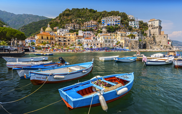 Bunte Boote schwimmen im Meer vor einer Küstenstadt mit Hügel und Gebäuden.