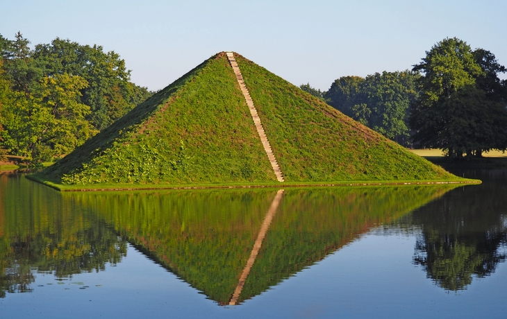 Eine grasbewachsene Pyramide spiegelt sich in einem ruhigen See.