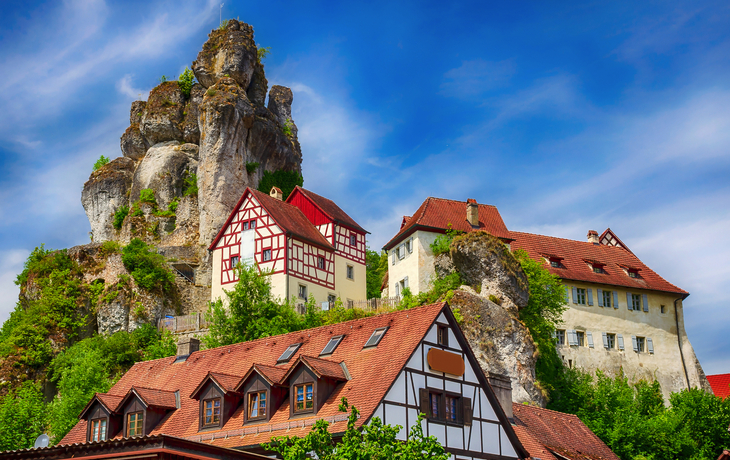 Fachwerkhäuser vor einer felsigen Anhöhe unter blauem Himmel.