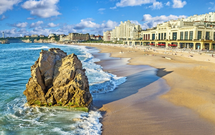 Strand mit Felsen, Stadtgebäude und blauem Himmel im Hintergrund.