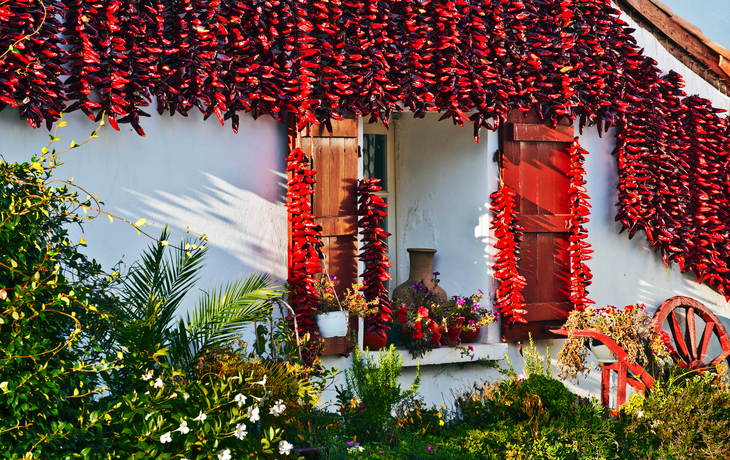 Weißes Haus mit roten Fensterläden und hängenden Chilischoten, umgeben von Pflanzen.