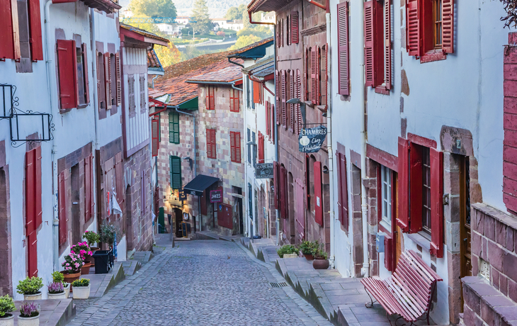Gepflasterte enge Straße mit traditionellen Häusern und roten Fensterläden.