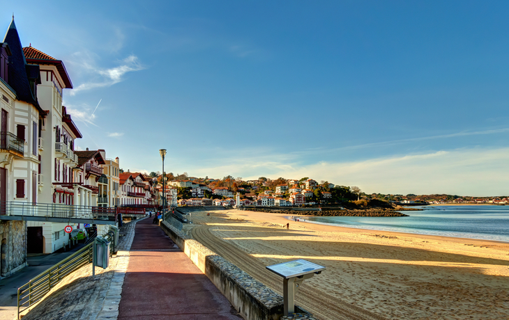 Küstenstraße mit Blick auf Sandstrand und Häuser an einem sonnigen Tag.