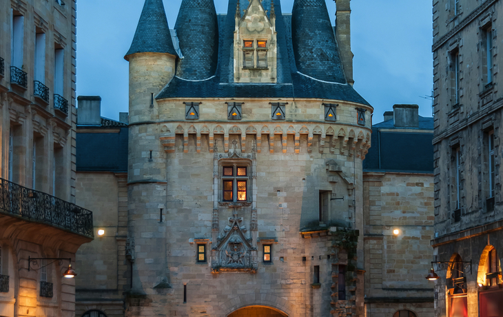 Abendstimmung vor der Porte Cailhau in Bordeaux, umgeben von Gebäuden und Straßenlaternen.