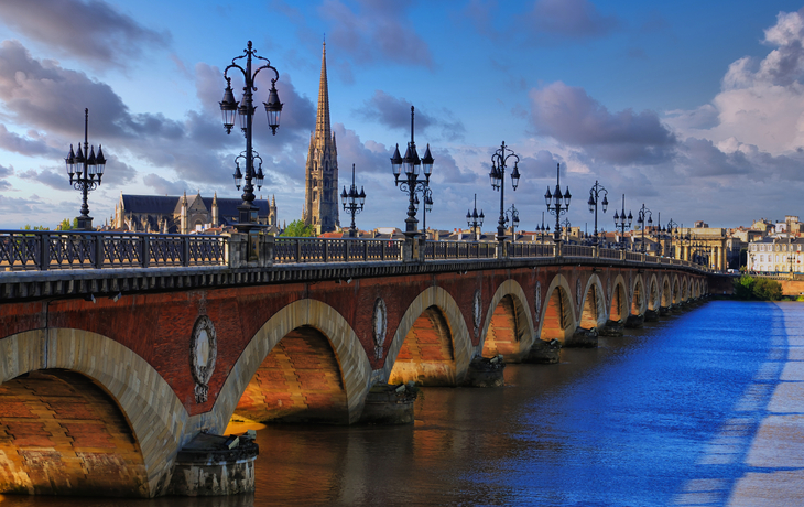 Historische Brücke über einen Fluss bei sonnigem Himmel mit Stadt im Hintergrund.