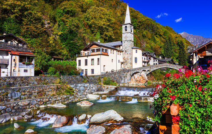 Kleines Dorf mit Kirche und Brücke über Fluss in einer Herbstlandschaft.