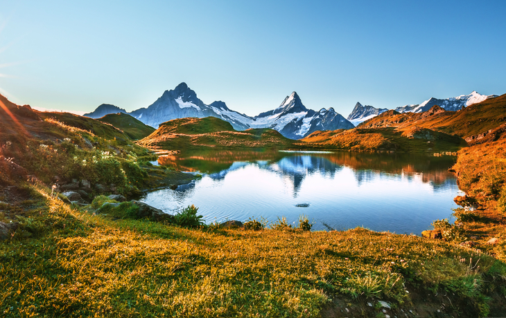 Berglandschaft mit See im Vordergrund und schneebedeckten Gipfeln im Hintergrund bei Sonnenaufgang.