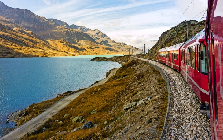 Ein roter Zug fährt entlang eines Sees mit Bergen im Hintergrund unter einem blauen Himmel.