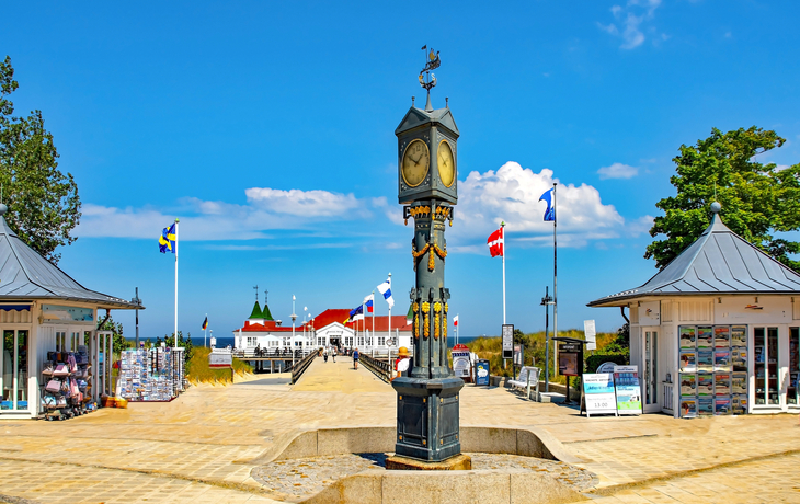 Uhr auf einer Promenade mit Pavillons und Flaggen bei sonnigem Wetter.