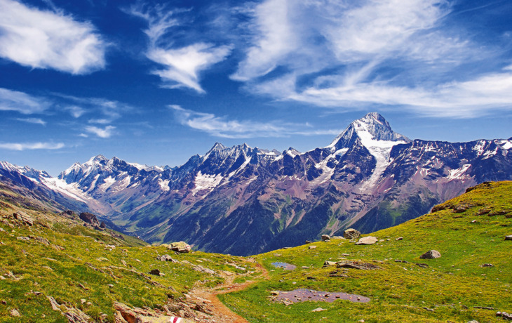 Berglandschaft mit Wiesen, schneebedeckten Gipfeln und blauem Himmel.