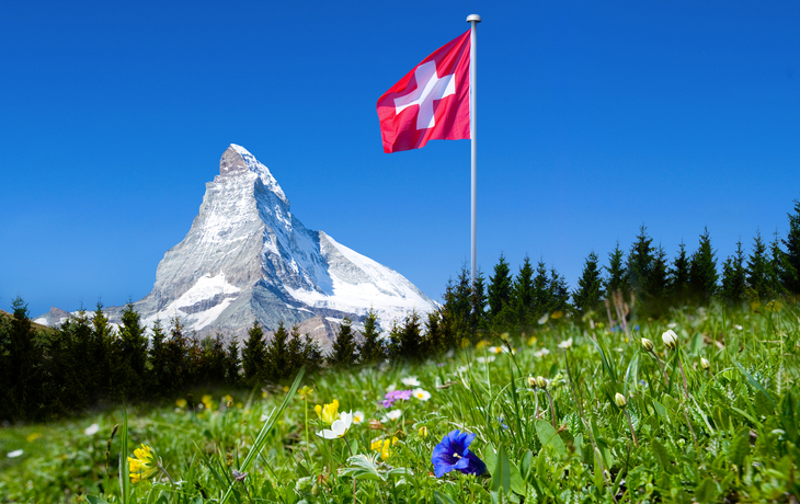 Schweizer Flagge vor dem Matterhorn und blühender Wiese.