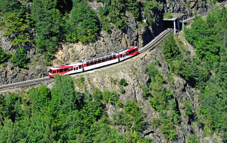 Rote Bergbahn fährt auf schmaler Gebirgstrasse durch bewaldetes Gebiet.