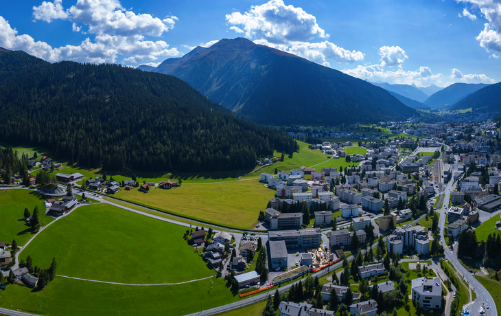 Panoramablick auf eine Stadt in einem Tal, umgeben von Bergen und grünen Wiesen.