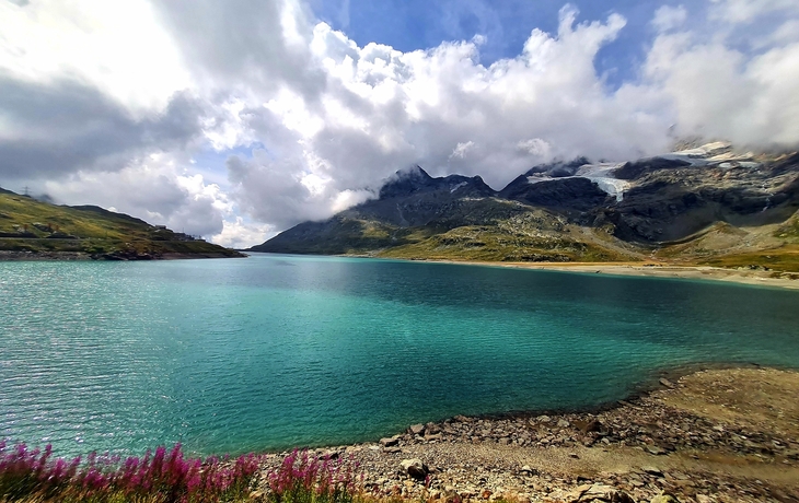 Bergsee mit türkisfarbenem Wasser und umliegenden Bergen unter bewölktem Himmel.