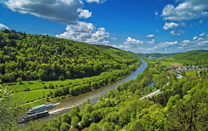 Fluss in grüner Hügellandschaft mit vorbeifahrendem Boot unter blauem Himmel.