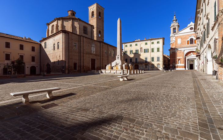 Weitläufiger, gepflasterter Platz mit Obelisk und historischen Gebäuden bei klarem Himmel.