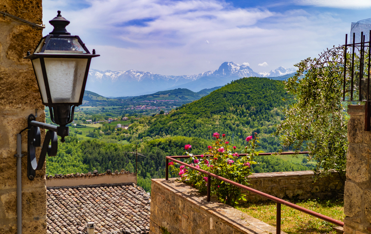 Blick auf grüne Hügel und Berge bei klarem Himmel, Steinmauer im Vordergrund.