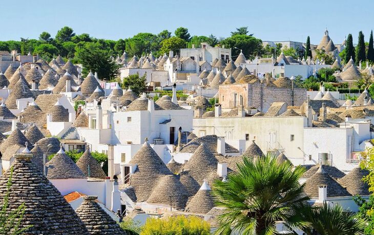 Weiß getünchte Trulli-Häuser mit Kegeldächern in einer Landschaft mit Bäumen.